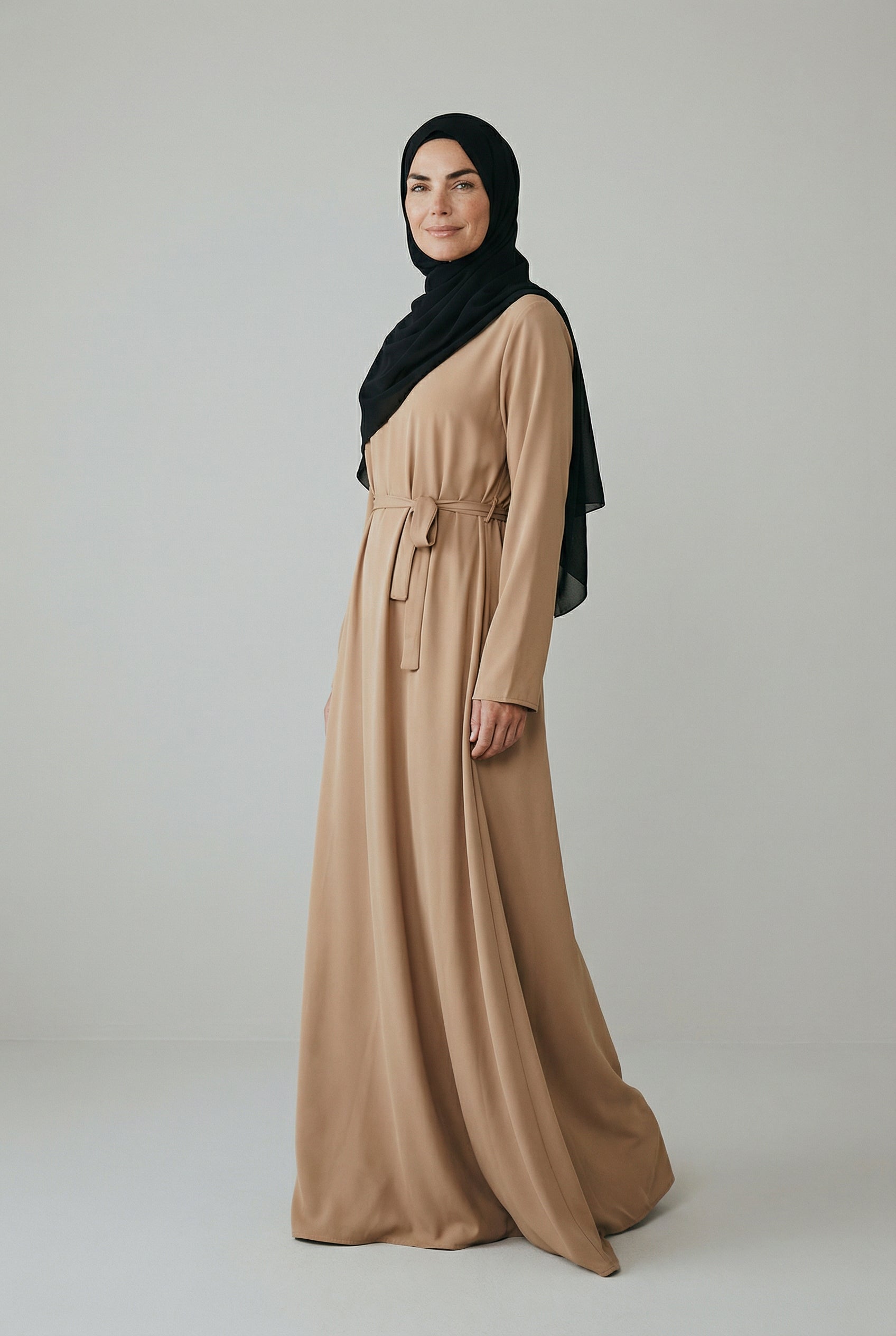 Woman wearing a long tan abaya and black hijab on a plain background